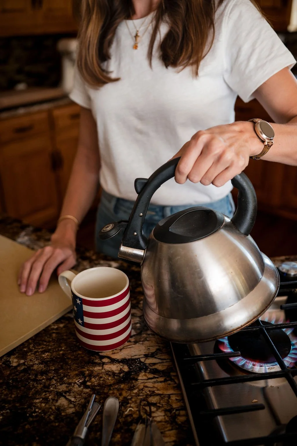 A woman in a white T-shirt and jeans is boiling water in a stainless steel kettle on a stove. The kettle is near a mug decorated with the American flag stripes and stars. The kitchen has wooden cabinets and a granite countertop.