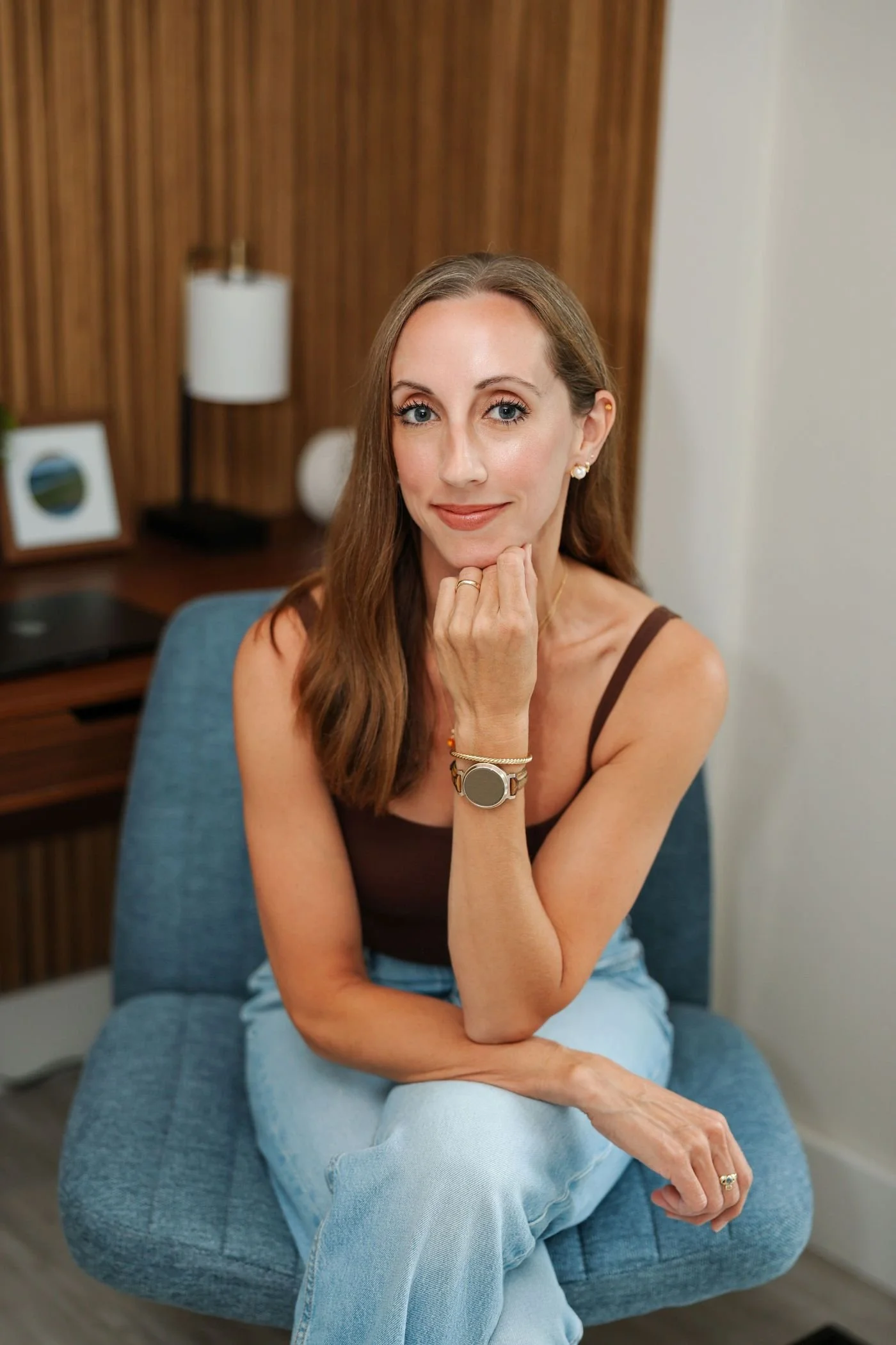 A woman with long brown hair, blue eyes, wearing a brown tank top and light blue jeans, sitting on a blue chair in an indoor setting with a wooden wall, a white lamp, and framed pictures in the background.