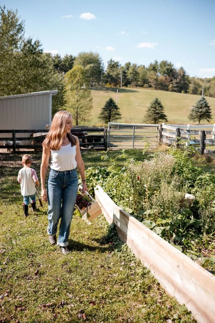 Jessica Nazzaro and a young girl walking along a garden bed filled with plants on a farm or rural property with green grass, trees, and a blue sky in the background.