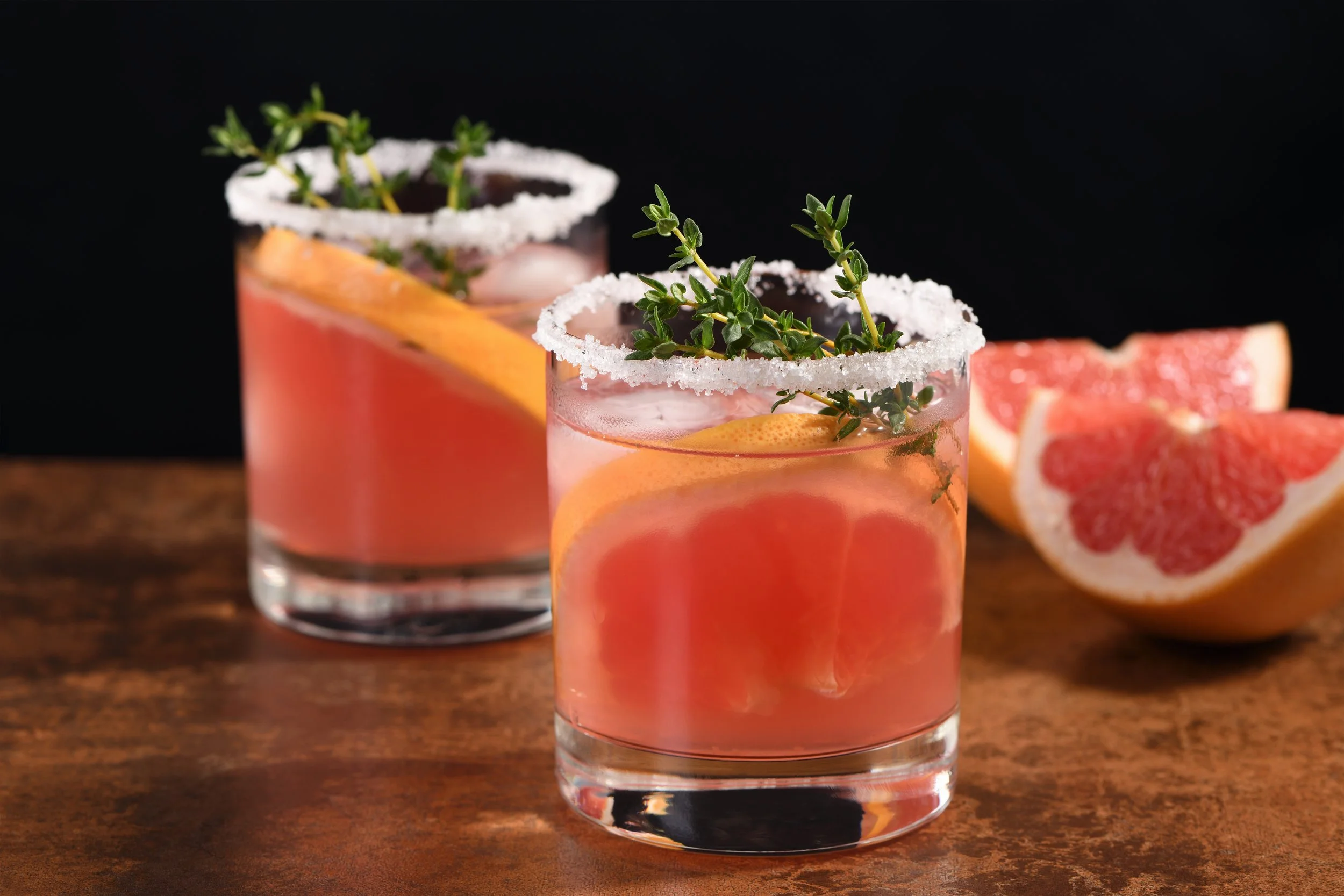 Two glasses of pink grapefruit cocktail garnished with fresh thyme, with salt-rimmed glasses, set on a rustic wooden surface, with grapefruit wedges in the background.