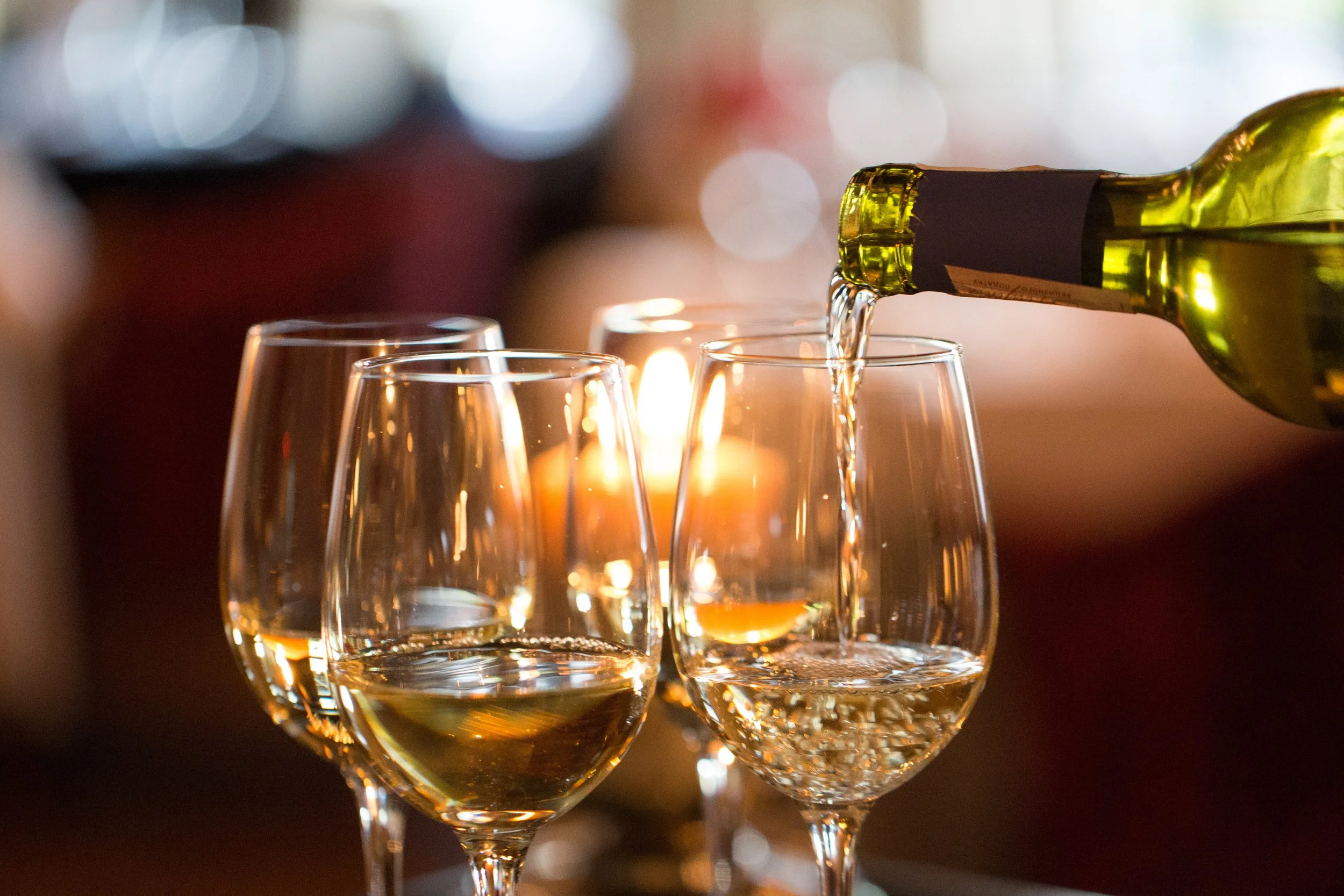 A green wine bottle is pouring white wine into a glass, with two other wine glasses filled with white wine in the background, against a warm, softly blurred background with candlelight.