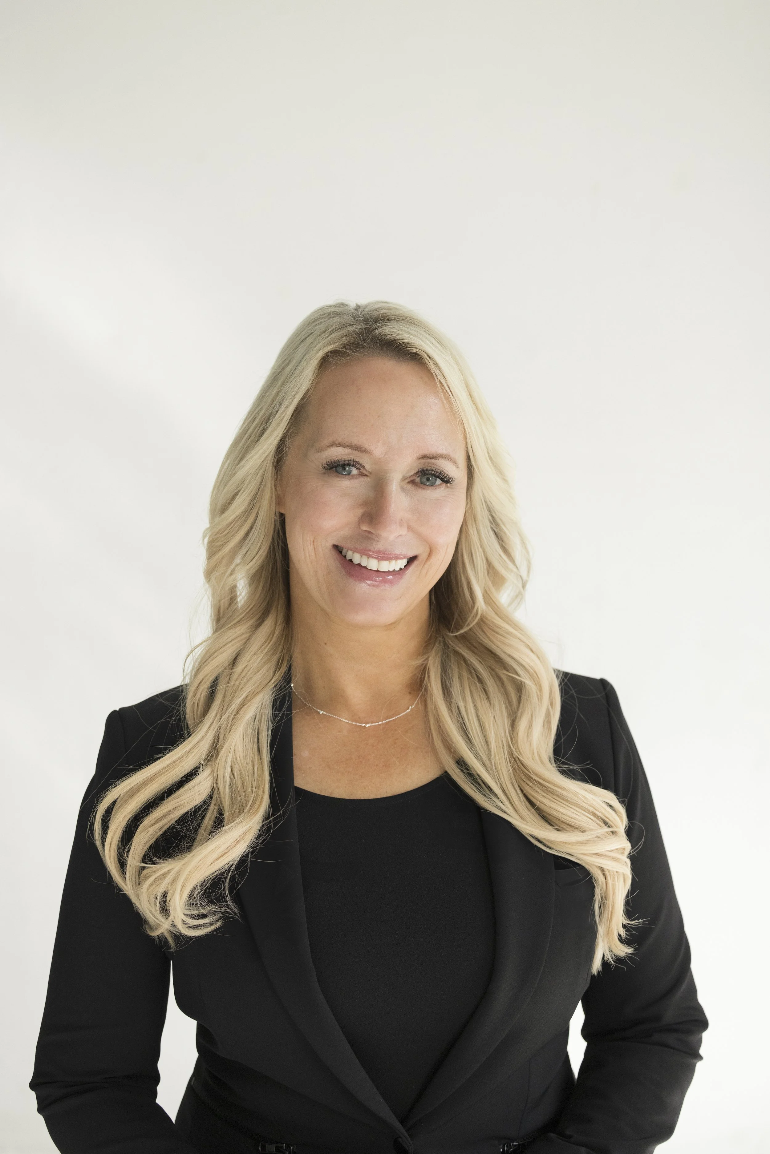 A woman with long blonde hair, wearing a black blazer and smiling at the camera, standing against a plain white background.