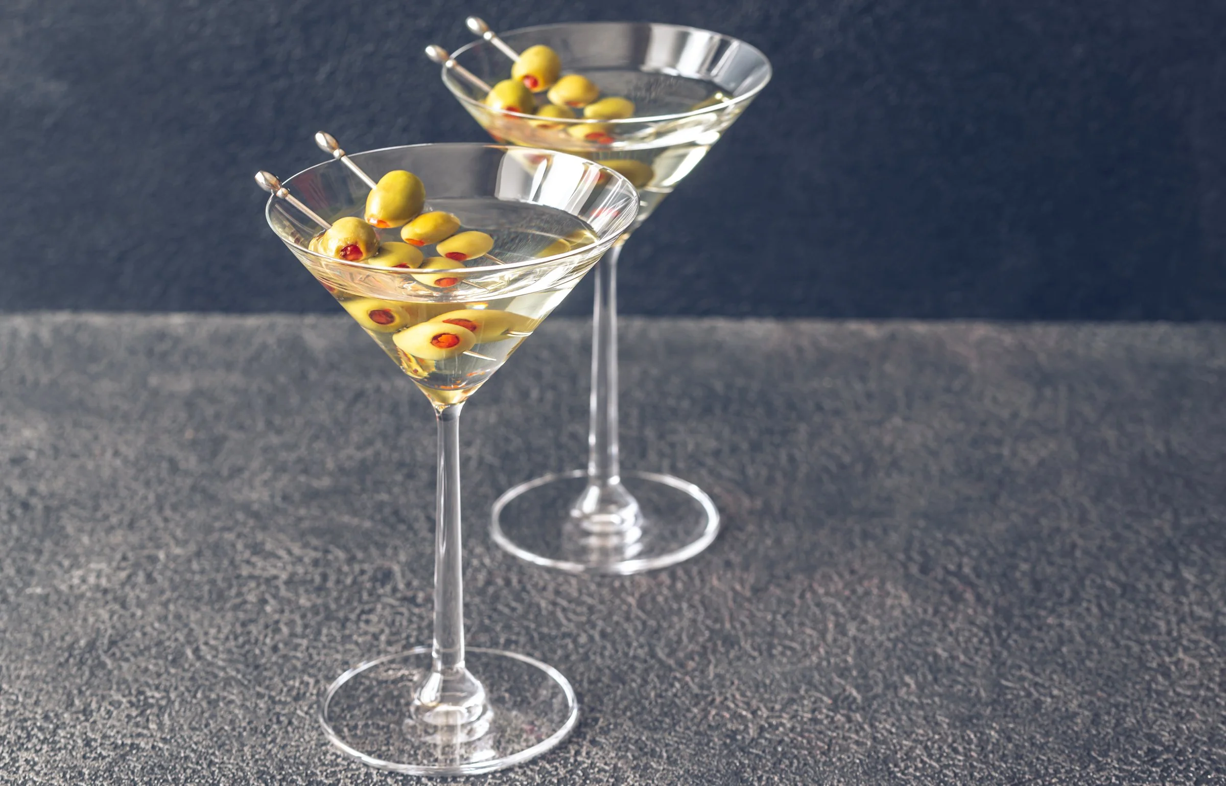 Two martini glasses filled with a clear liquid, each with green olives on toothpicks, on a dark textured surface.