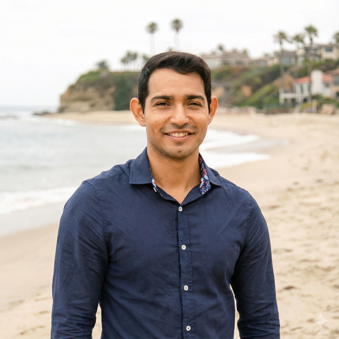 A smiling man in a dark blue button-up shirt standing on a beach with ocean waves, sandy shore, and residential houses with palm trees in the background.
