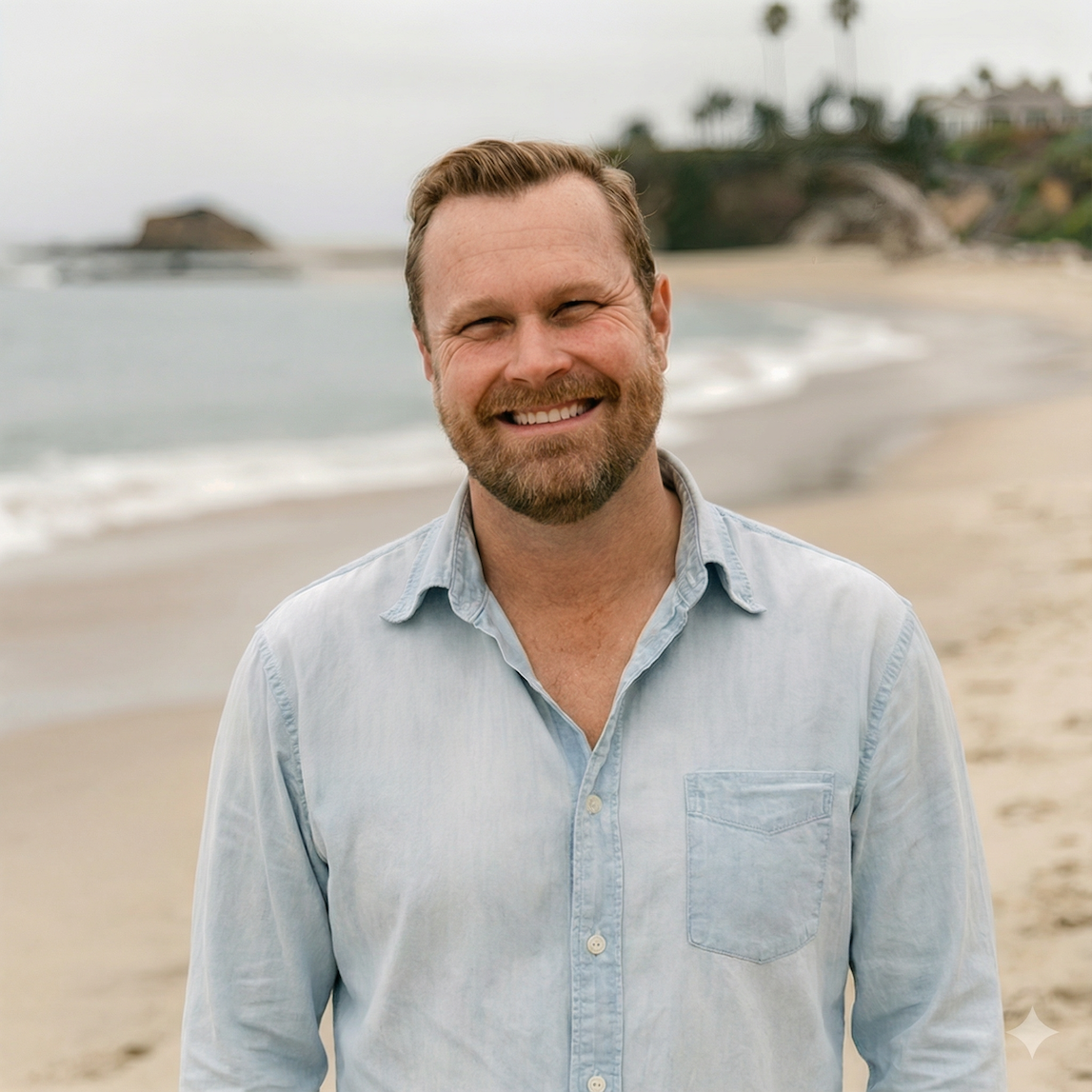 Smiling man with a beard in a light blue shirt standing on the beach with ocean and cliffs in the background.