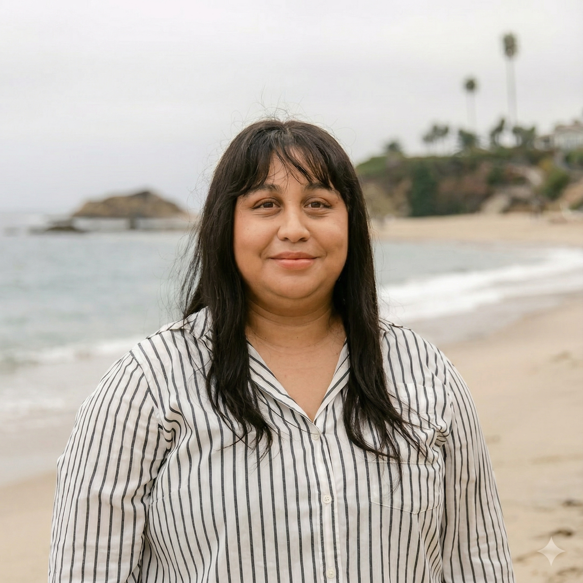Woman with dark hair and light skin standing on a beach with ocean and cliffs in the background.