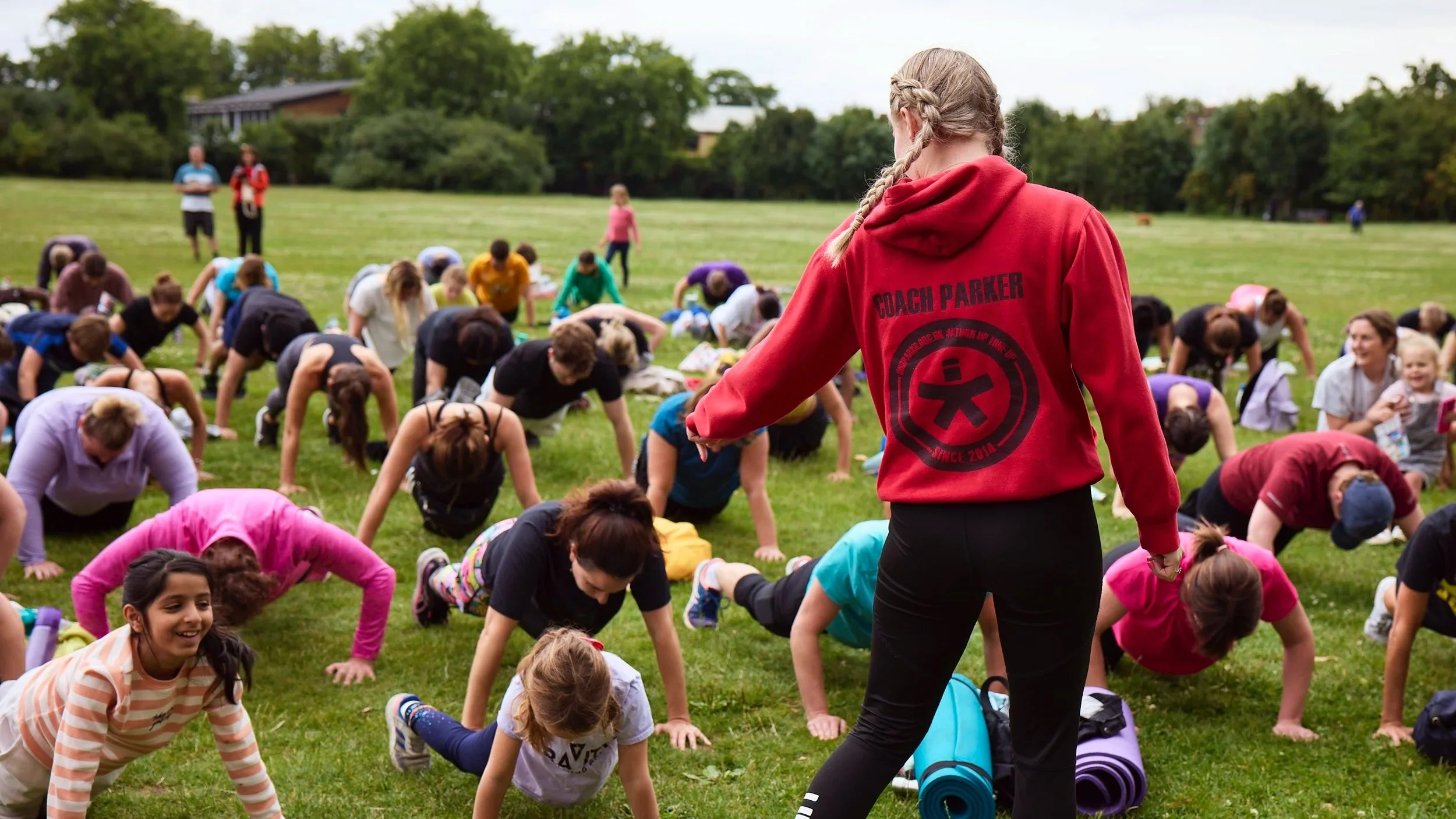 A group of children participating in an outdoor yoga class on a grassy field, led by an instructor wearing a red hoodie with 'Coach Parker' written on the back.