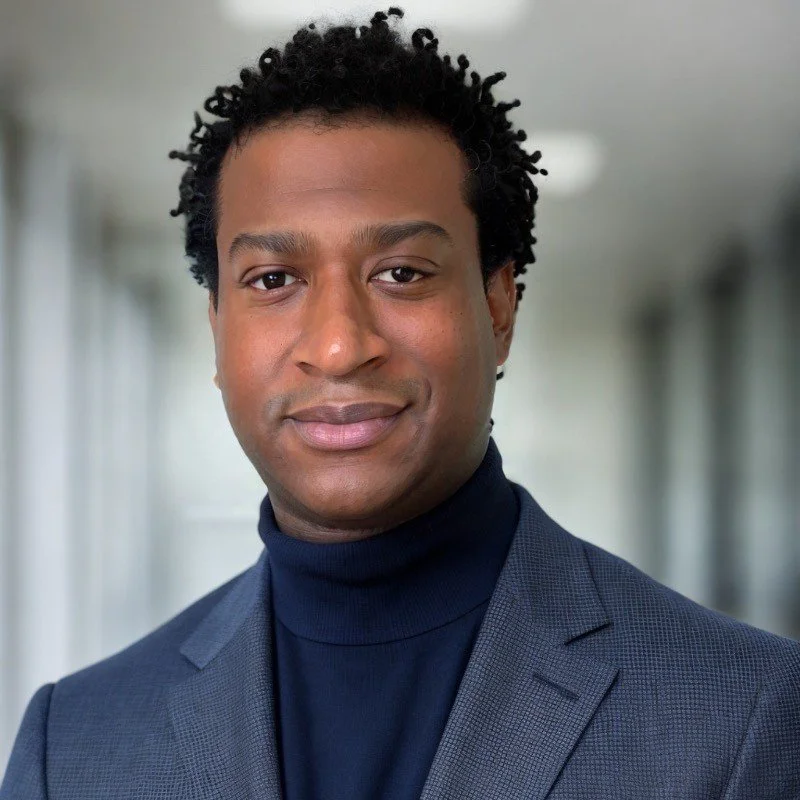 Headshot of a man in a dark blazer and turtleneck, with short curly hair, smiling slightly, professional indoor background.