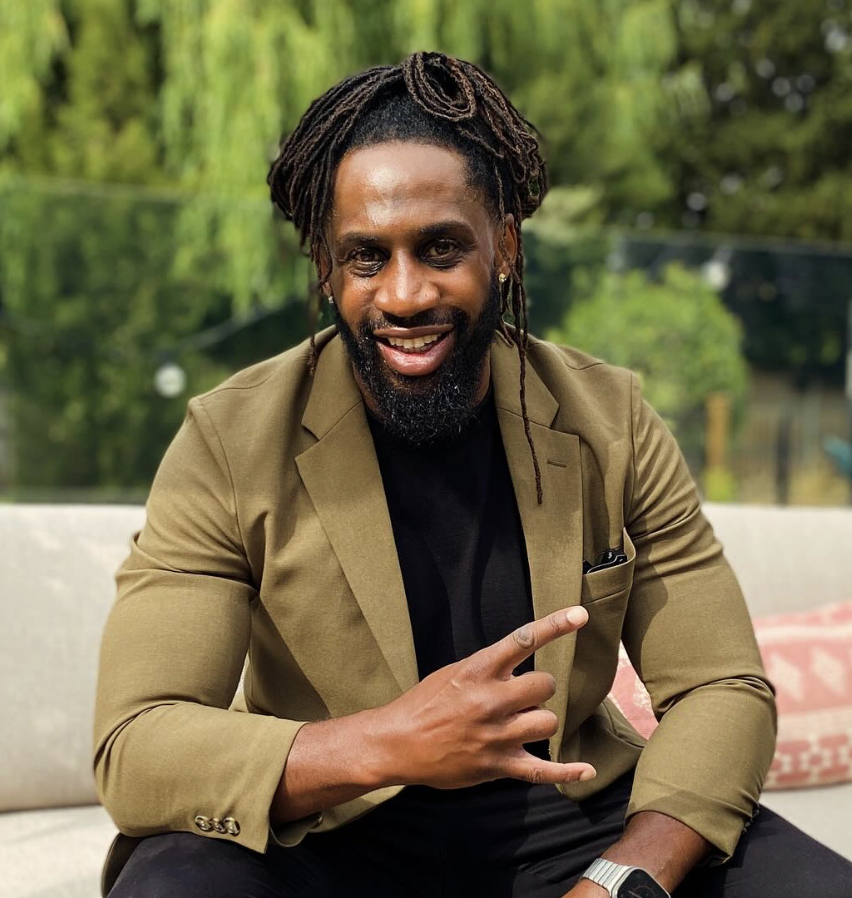 A man with braided hair, a beard, and mustache, smiling and gesturing with his right hand in a peace sign. He's wearing a khaki blazer over a black shirt, sitting outdoors with green trees in the background.
