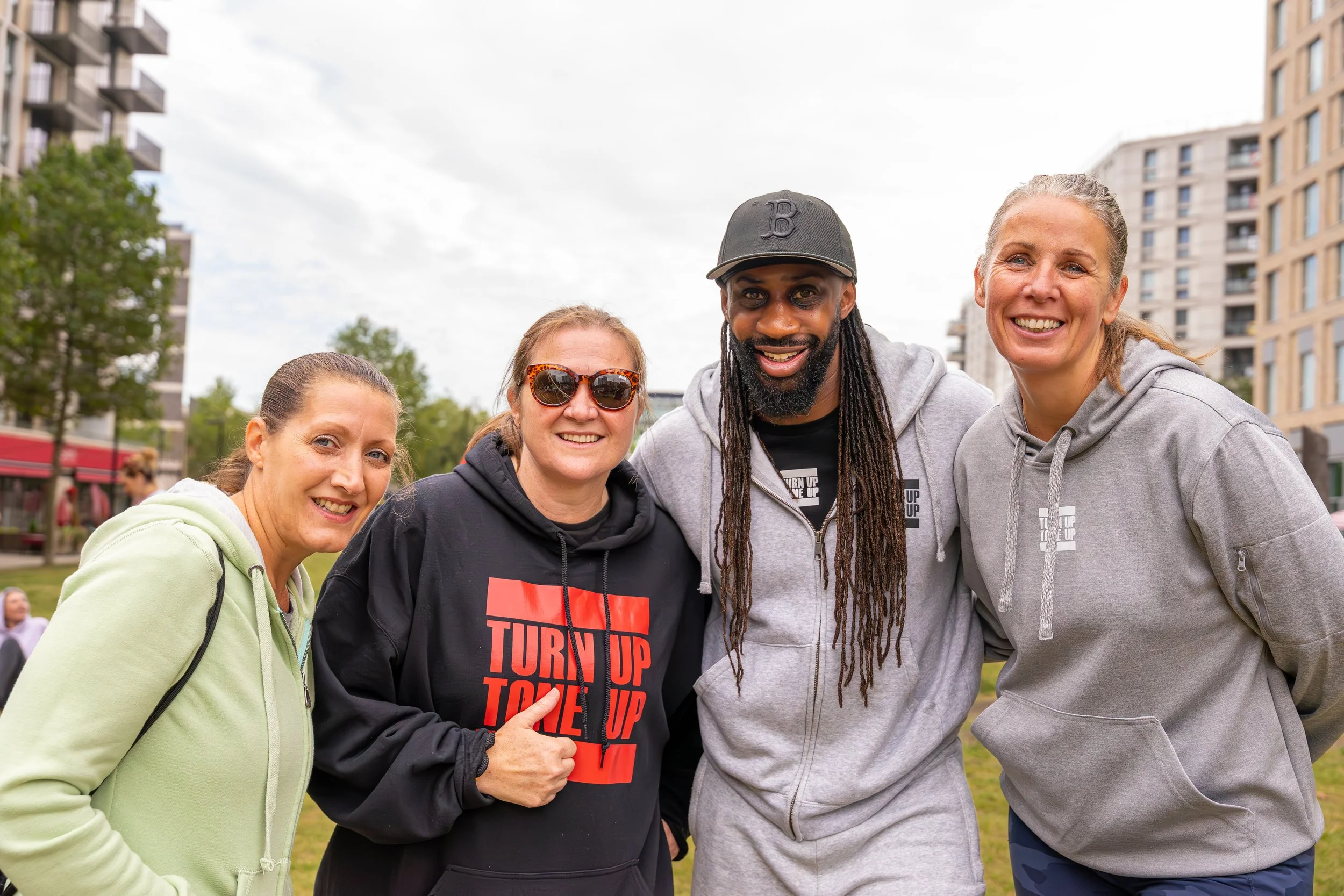 Group of four diverse people smiling and posing outdoors in a park during a community event, some wearing hoodies with motivational messages, in front of apartment buildings with trees and cloudy sky.