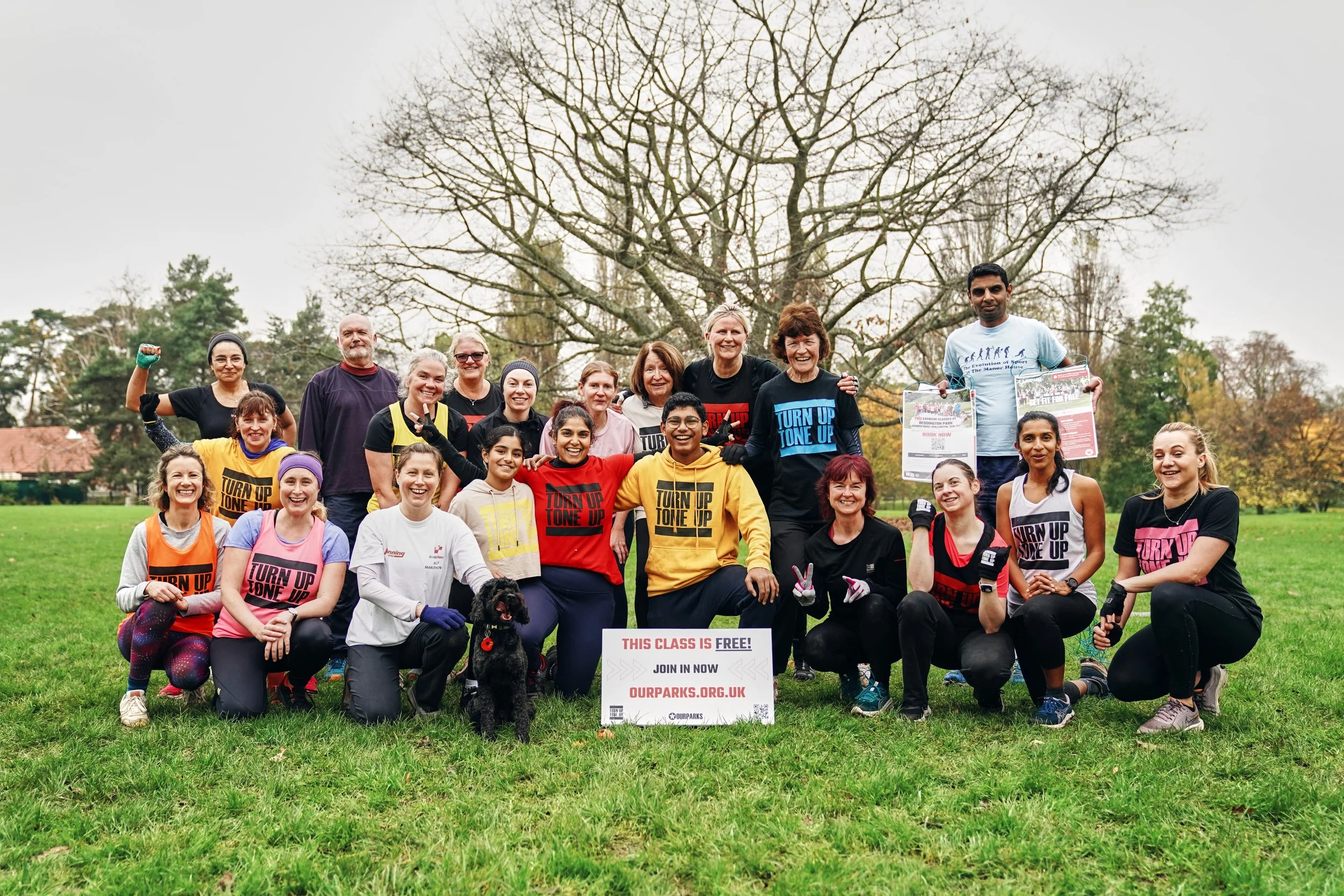 Group of diverse women and a few men participating in an outdoor fitness event called 'Turn Up Tone Up', posing with smiles and gestures in a park with grass and trees, some wearing exercise shirts, holding signs, and accompanied by a black dog.