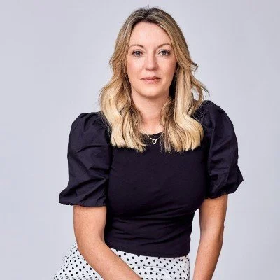 A woman with blonde hair in a black puff-sleeve top and a polka dot skirt, posing against a light gray background.