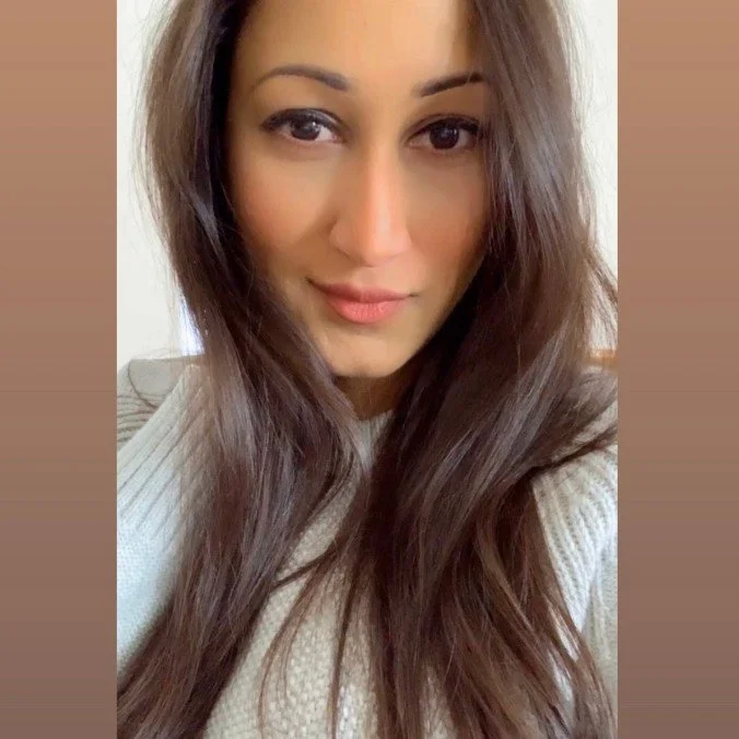 A young woman with long, wavy brown hair and light makeup, wearing a beige sweater and smiling at the camera.
