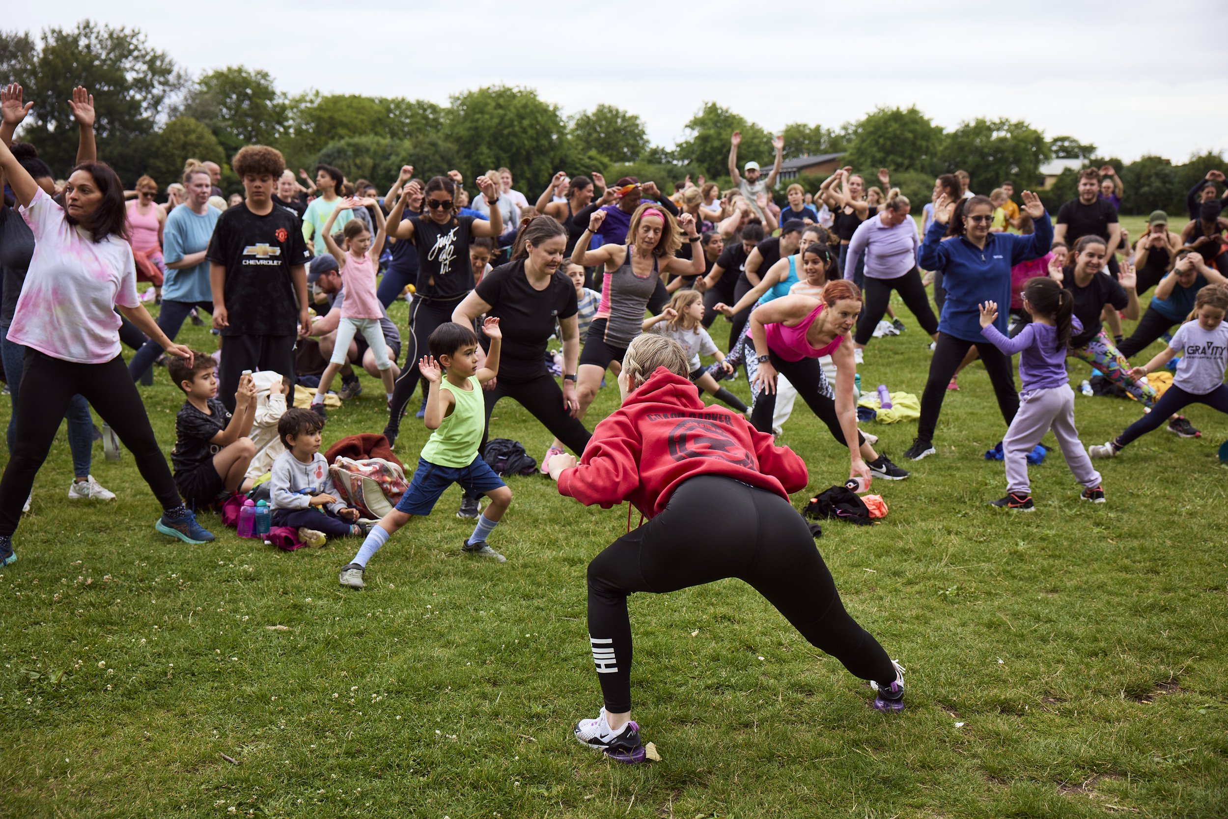 Groups of people participating in an outdoor fitness class on a grassy field, with children and adults stretching and exercising under cloudy skies.