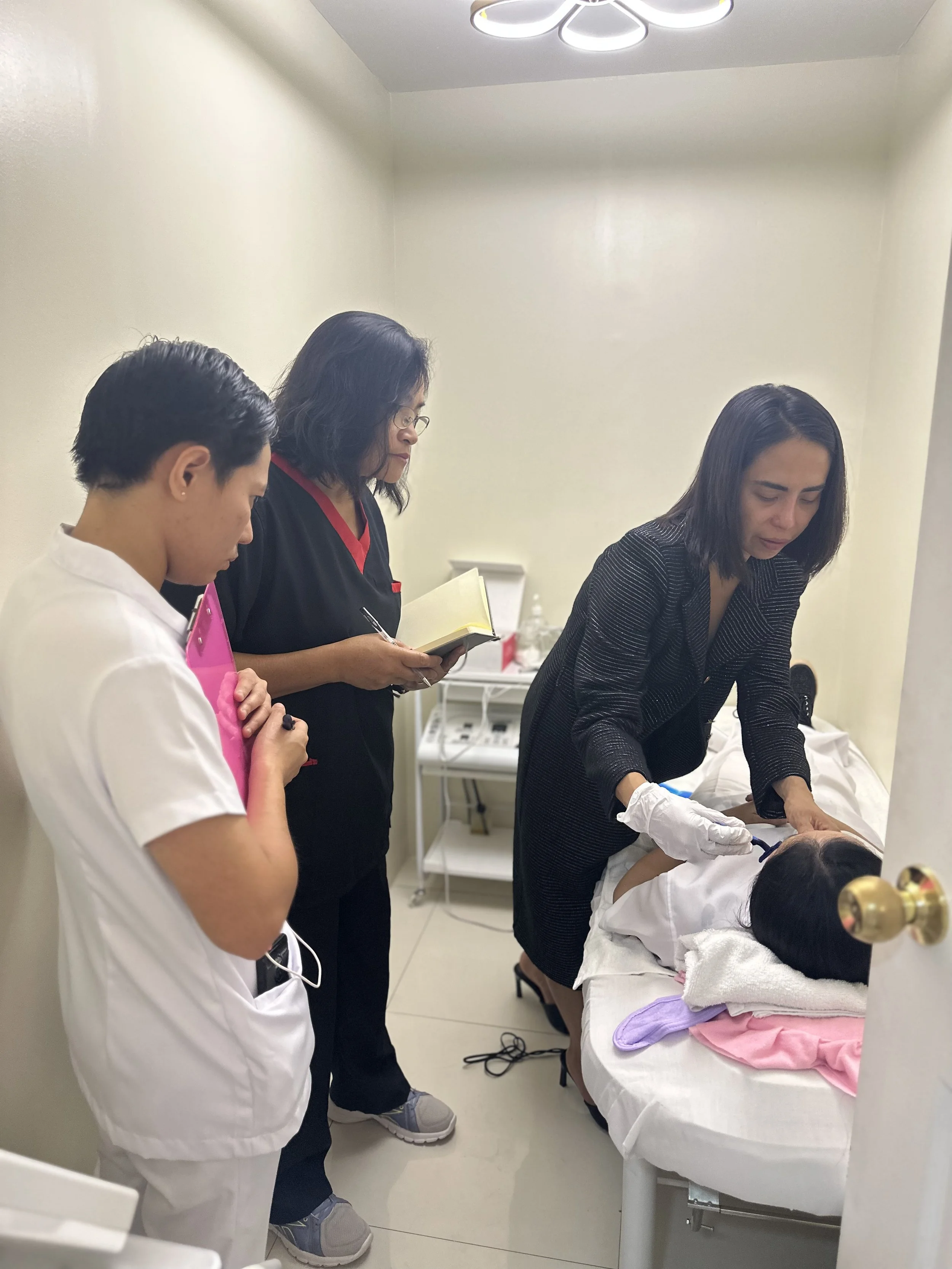 Female doctor in black attire attending to a patient lying on a hospital bed, with two healthcare professionals observing, one of whom is taking notes