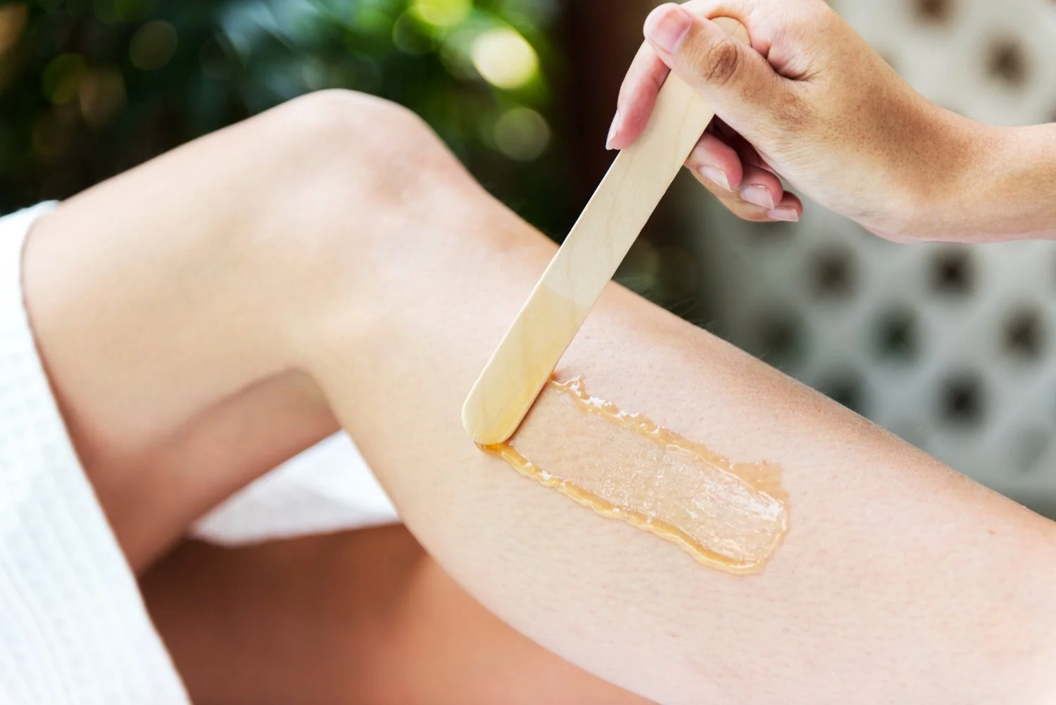 A person applies wax to their leg using a wooden spatula during a waxing treatment, with a blurred background of outdoor greenery.