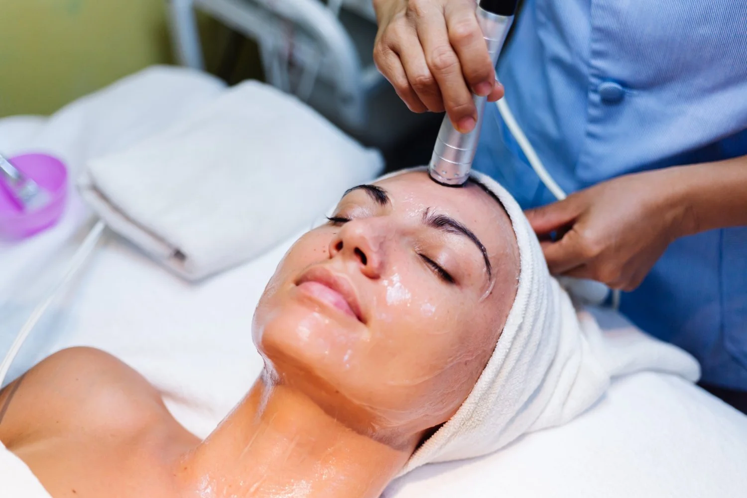 A woman receiving a facial treatment with a device on her forehead, lying with her eyes closed, in a spa setting.