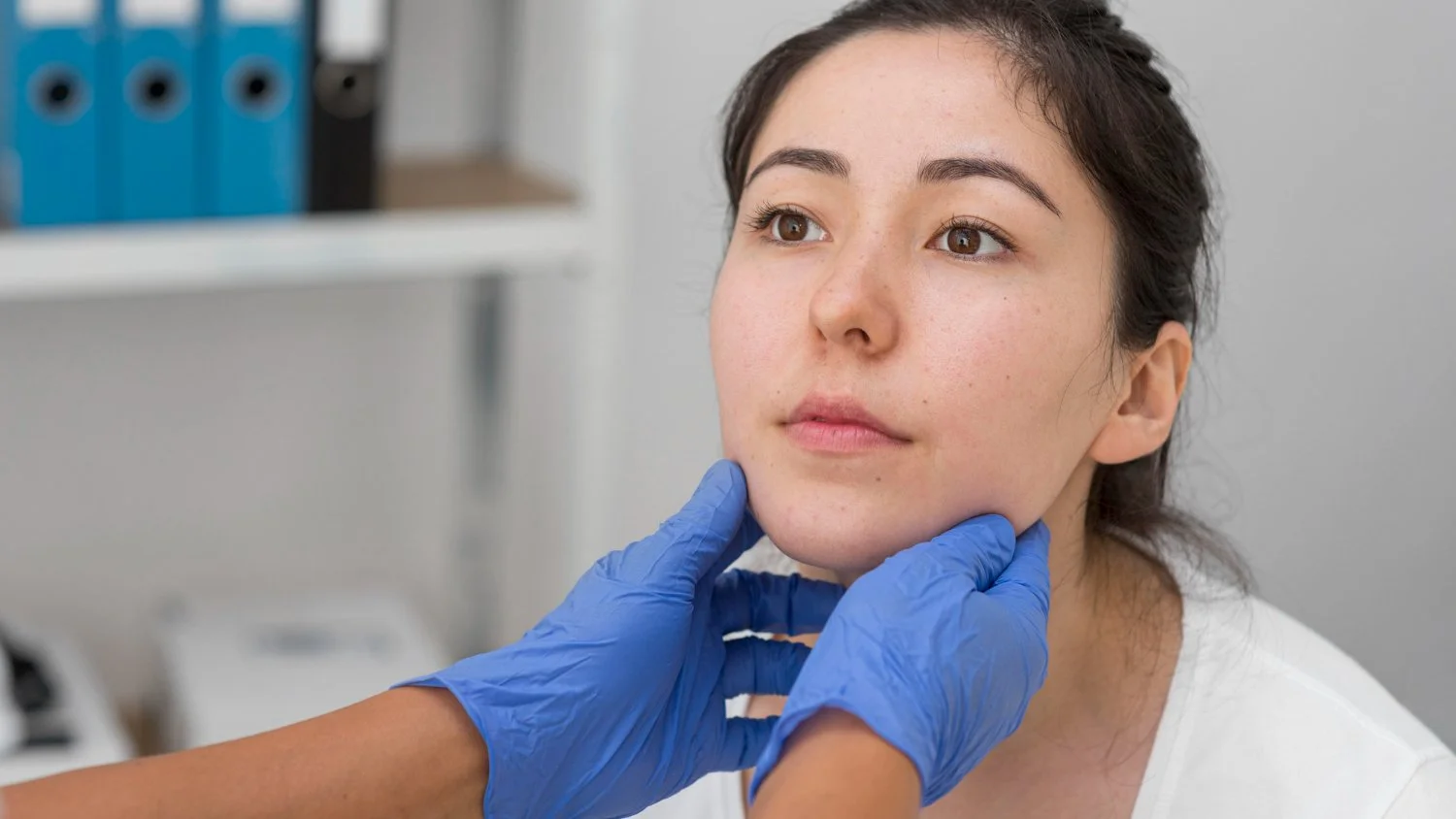 A woman getting her face examined by a healthcare professional wearing blue gloves, likely at a medical facility.