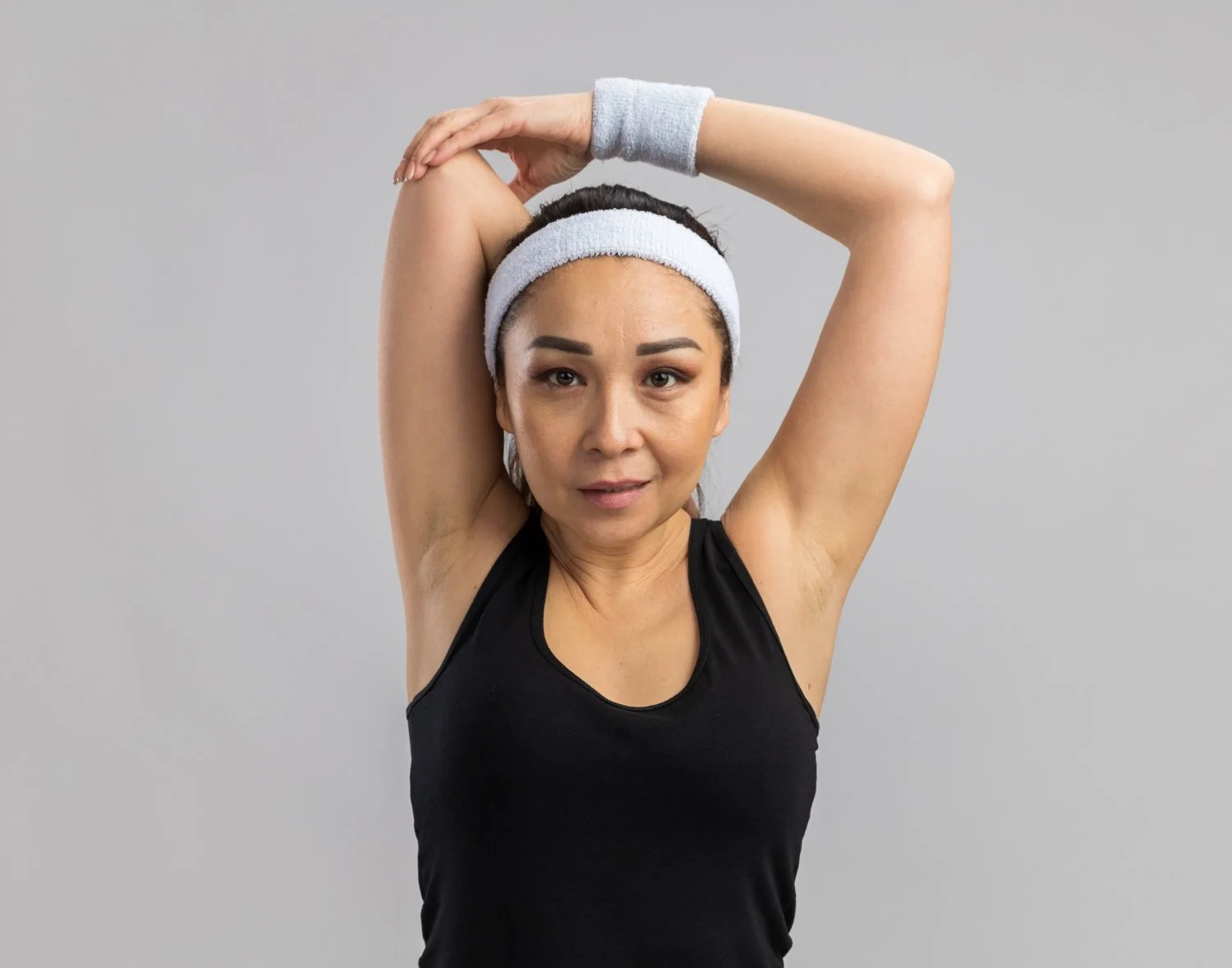 Woman in black tank top with white headband and wristband stretching her right arm over her head against a plain gray background.