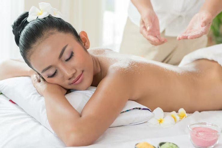 A woman receives a massage in a spa setting, lying face down on a massage table with her eyes closed. A spa therapist is applying massage oil or lotion on her back, with some flowers placed nearby.