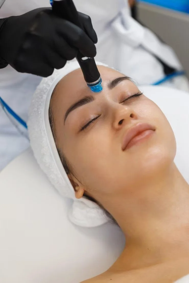 A woman lying in a spa or clinic bed, receiving a skincare treatment on her forehead from a professional wearing black gloves, with her eyes closed and relaxed.