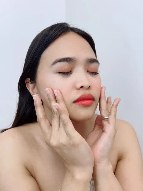 A woman with long dark hair and makeup, touching her face gently with her eyes closed, standing against a plain background.
