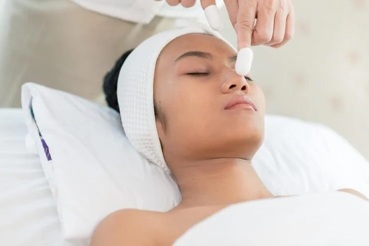 Person lying in bed with eyes closed, receiving a facial treatment with a cotton pad in a clinical setting.