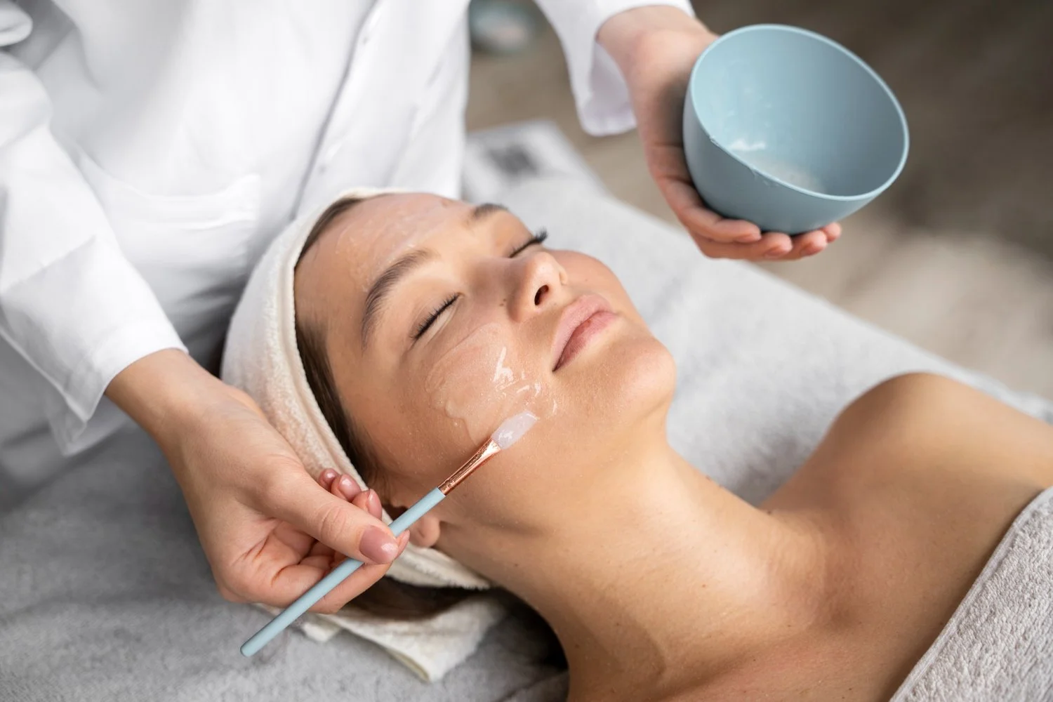 A woman receiving a facial treatment with cream applied on her face using a brush, while lying on a treatment bed with a towel wrapped around her head.