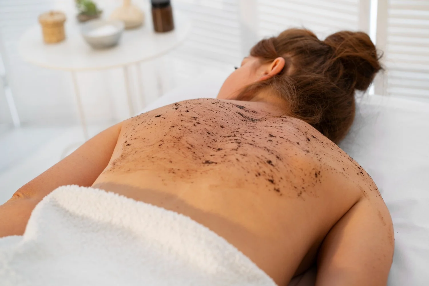 A woman lying face down on a massage table with a face pillow, covered with a towel around her waist, receiving a body scrub with coffee grounds on her back in a spa room.