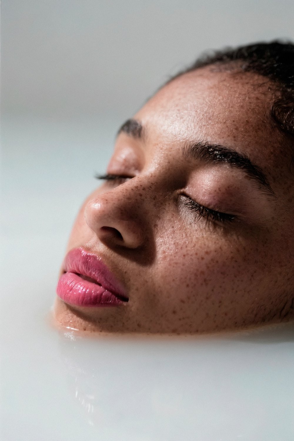Close-up of a woman's face with closed eyes, partially submerged in water, displaying her skin texture and freckles.