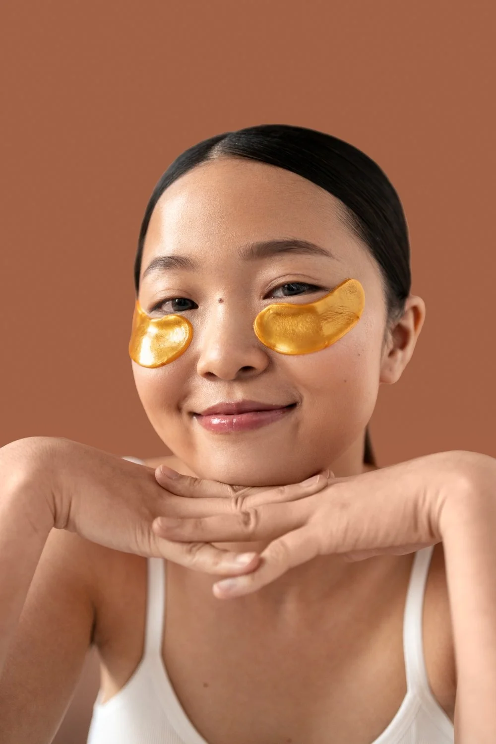 A woman with dark hair slicked back, wearing white tank top, relaxing with golden under-eye patches, resting her chin on her hands, smiling softly against a brown background.