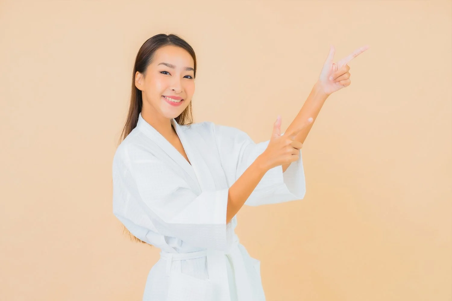 A smiling woman in a white martial arts uniform pointing to her right against a beige background.