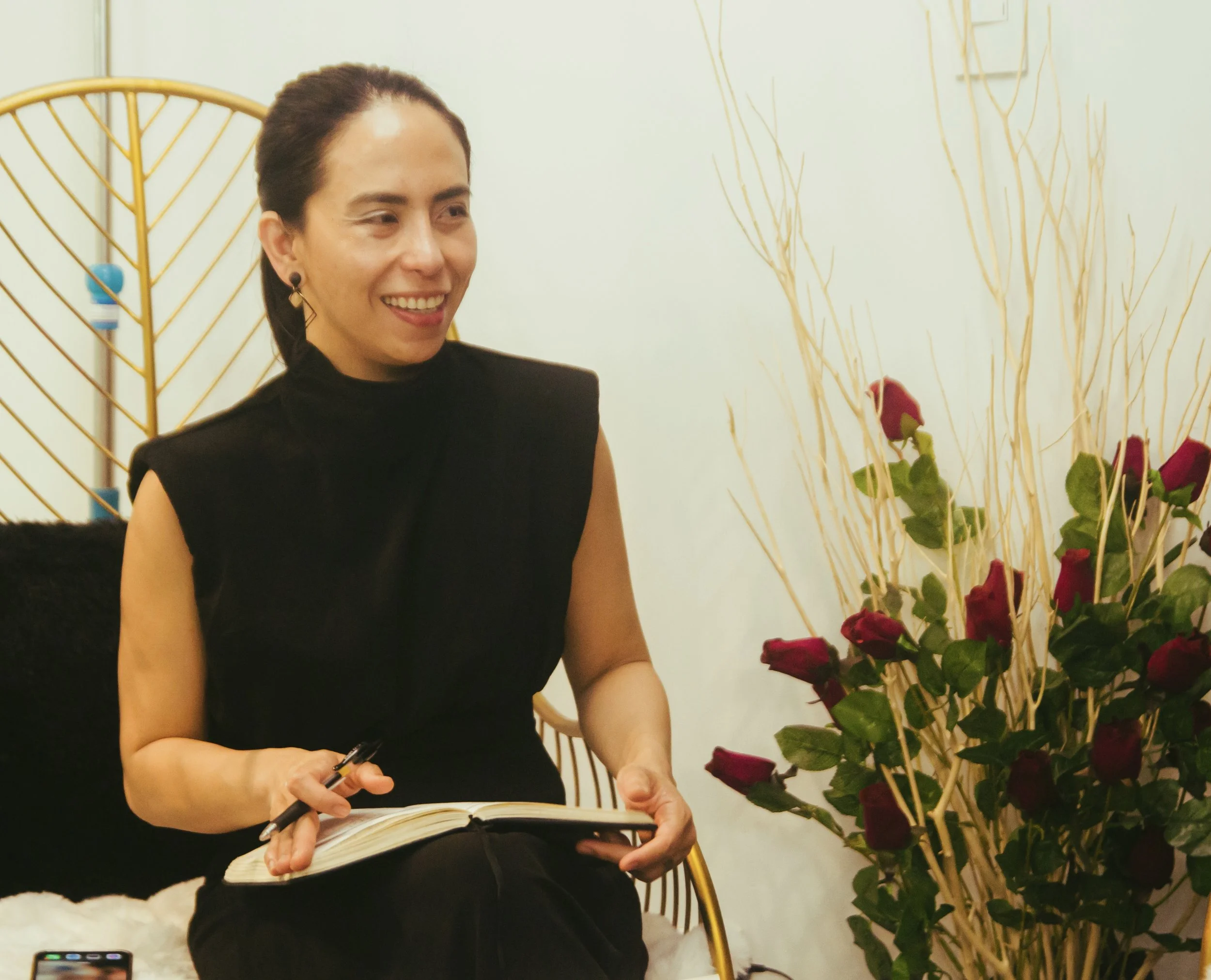 A woman with dark hair pulled back, wearing a black sleeveless top, sitting indoors with a notebook and pen, smiling. There are red roses and decorative dry branches beside her, and a white wall in the background.