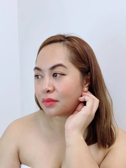 A woman with light skin and reddish-brown hair looking to the side, touching her ear, against a plain white wall.