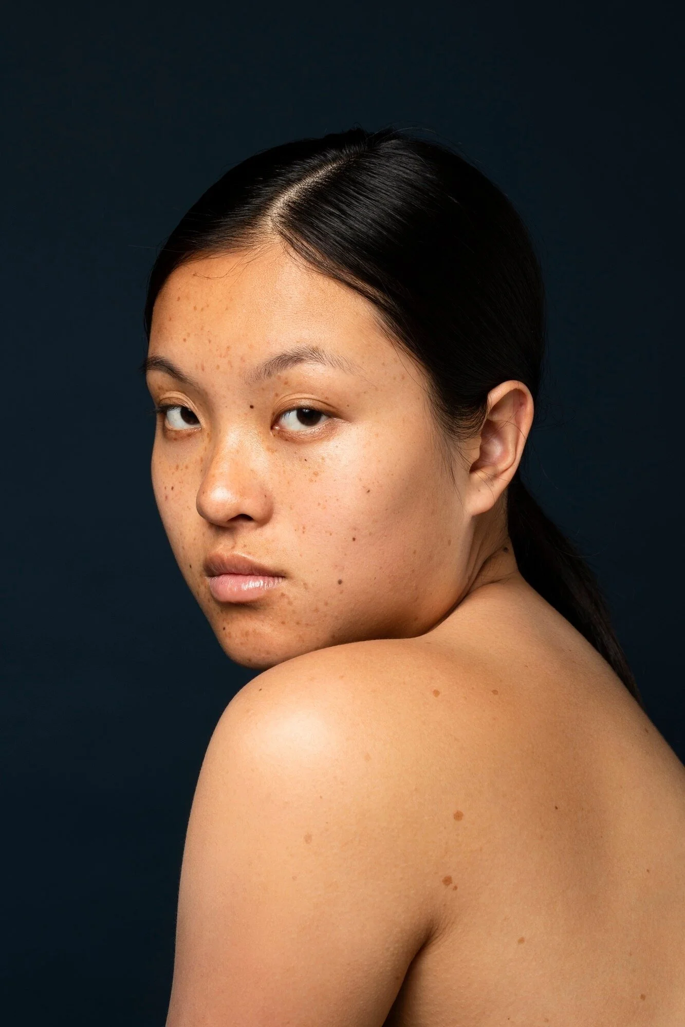 Close-up portrait of a woman with dark hair and visible freckles on her face and shoulder, looking over her shoulder with a neutral expression against a dark background.
