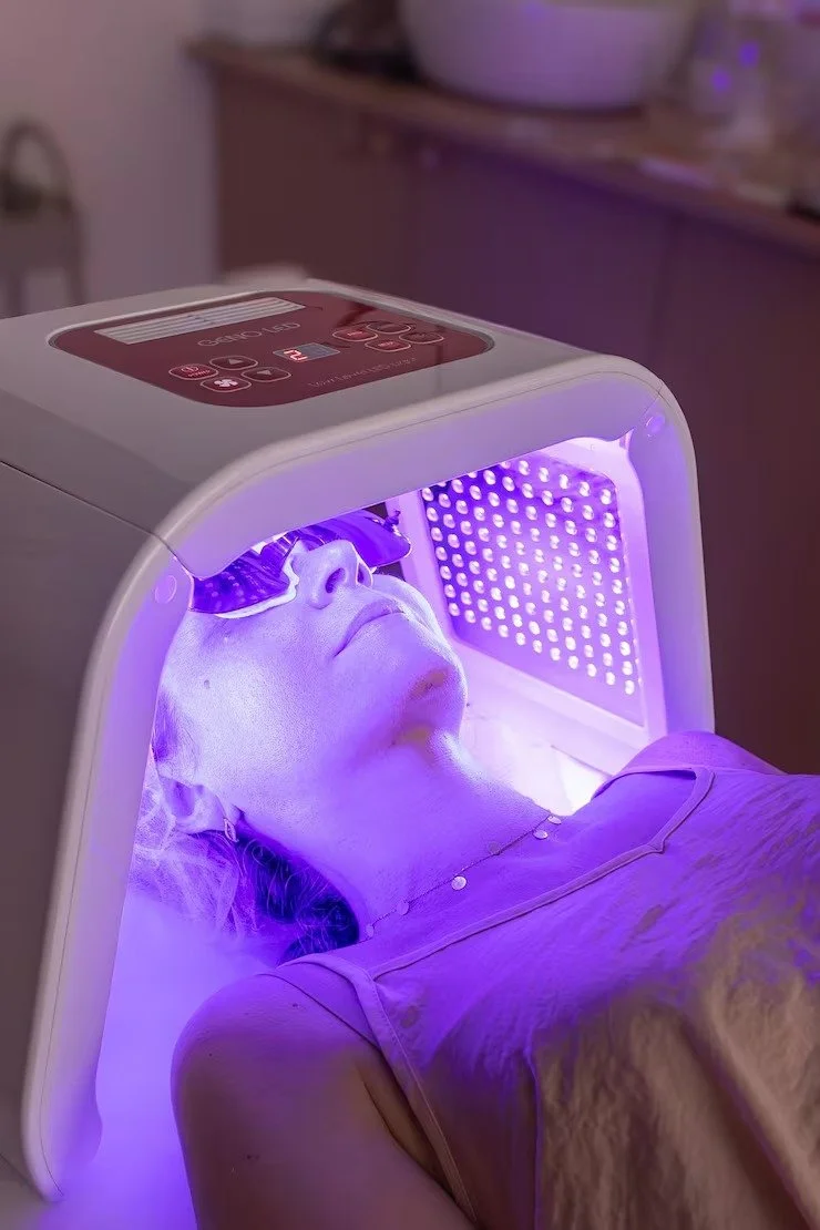 Woman lying under a red and white LED light therapy device with purple UV light, wearing protective glasses, in a spa or wellness setting.