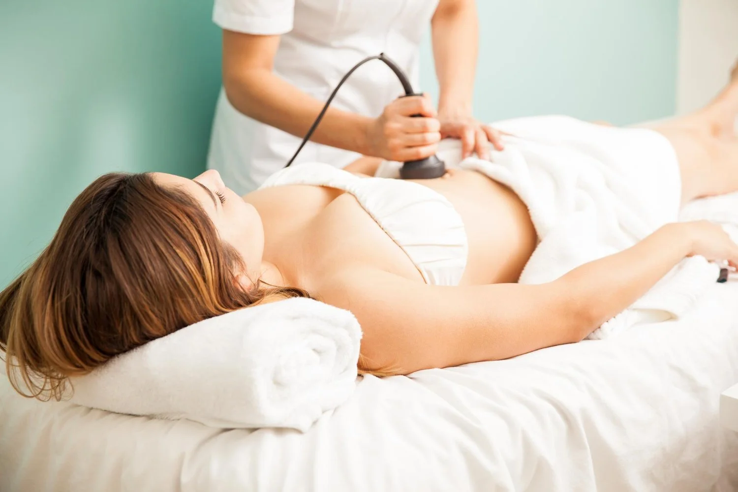 A woman lying on a treatment table with eyes closed, receiving a body scan or ultrasound from a healthcare professional in a clinical setting.
