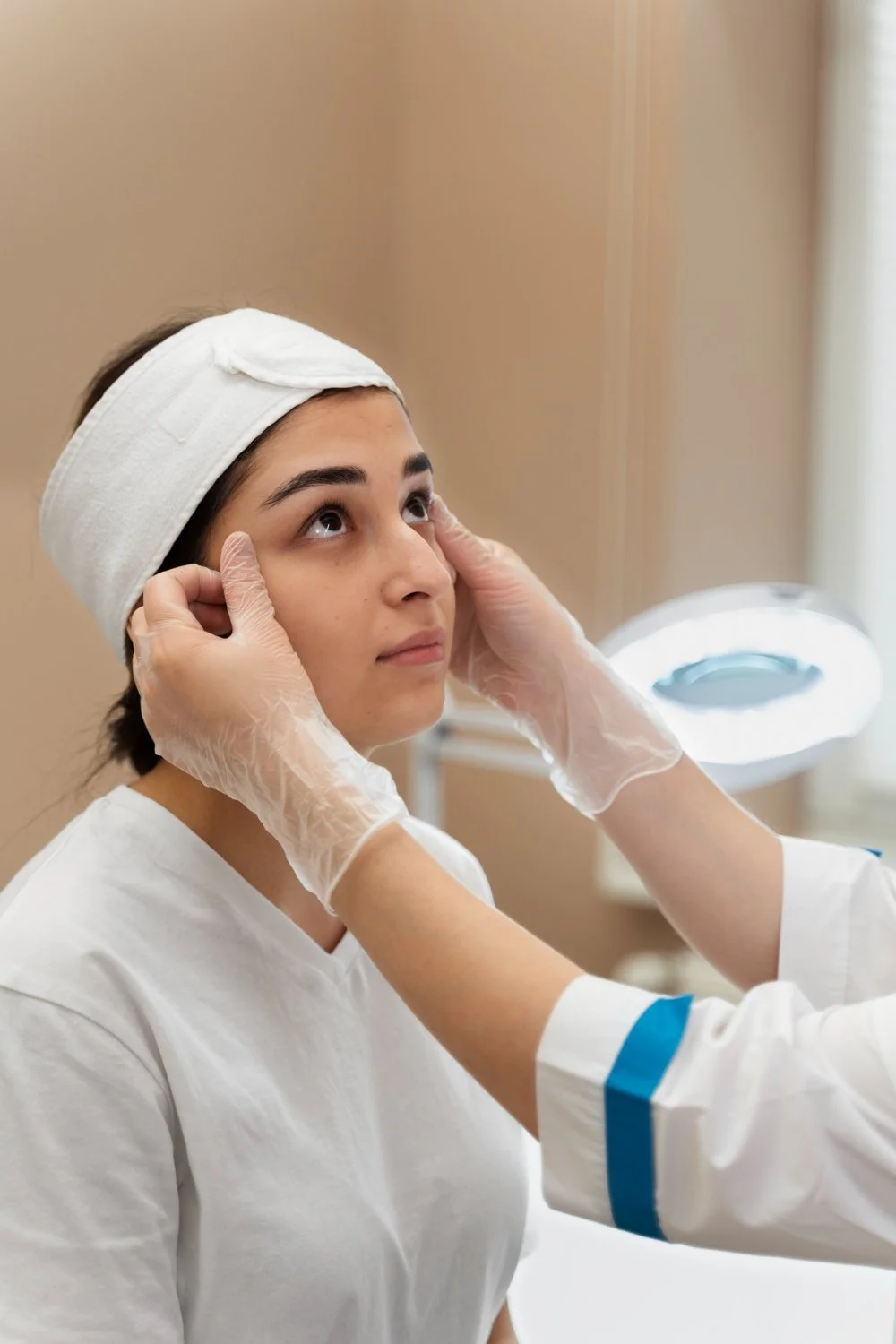 A woman wearing a white headband having her face examined by a medical professional in a clinical setting.