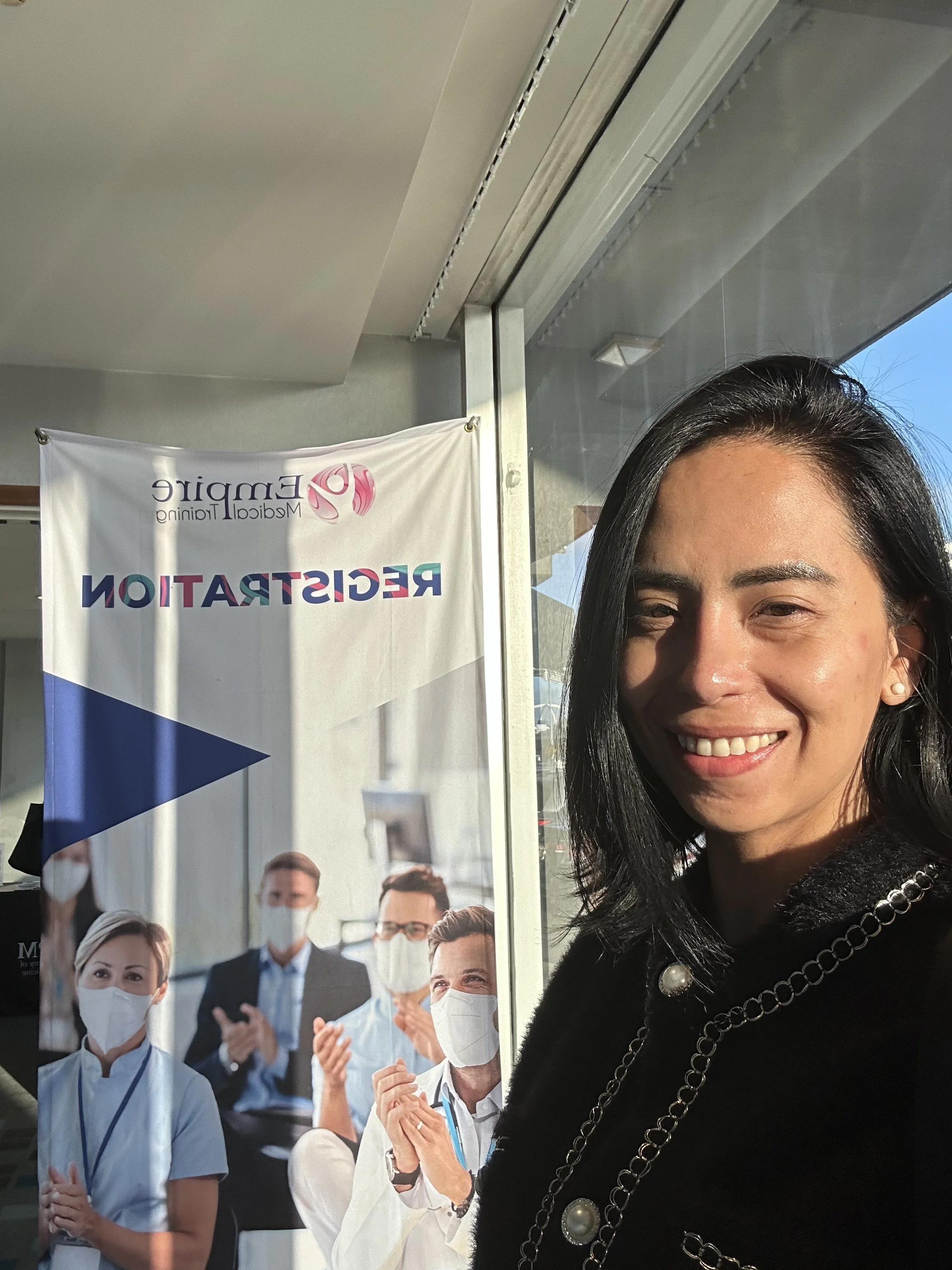 A woman taking a selfie indoors with a banner in the background that reads 'Reception' and features images of healthcare professionals wearing masks.