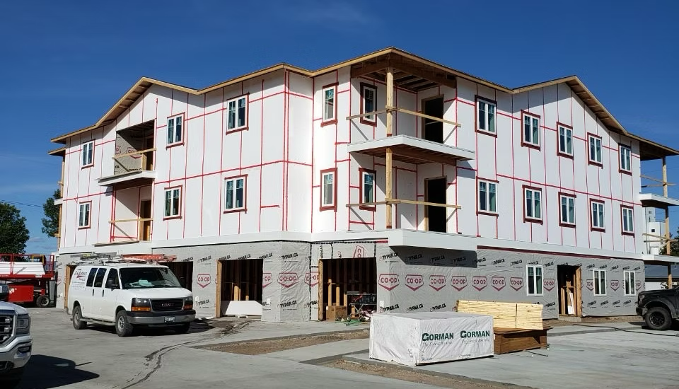 Four-story building under construction with framed exterior walls, construction materials in front, and several vehicles parked nearby.