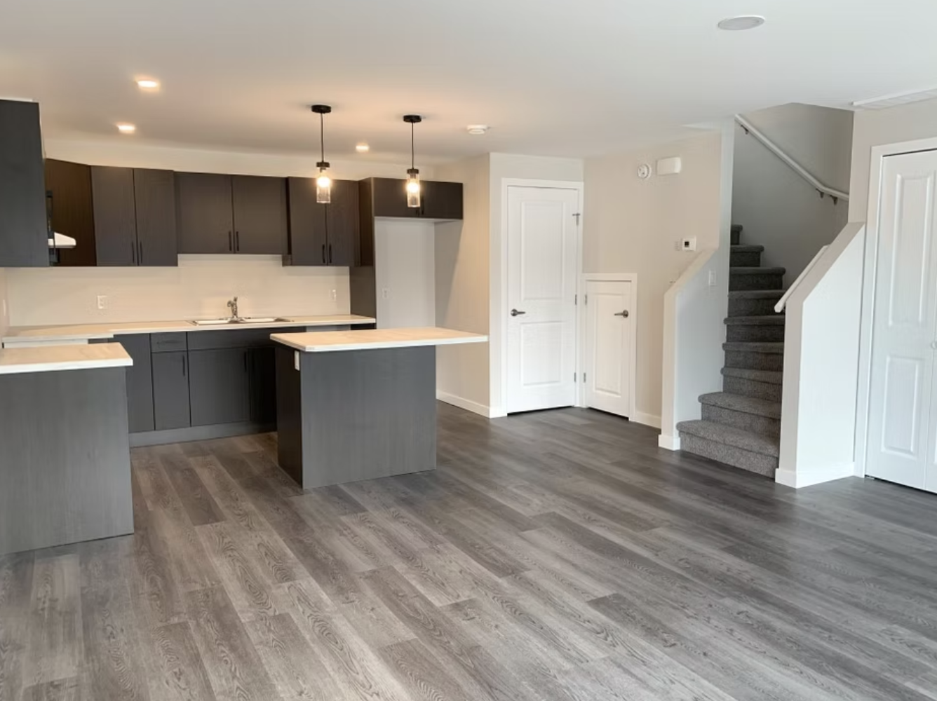 Open-concept kitchen with dark gray cabinets, light wood countertops, and a small island. The space features gray hardwood flooring, white walls, a staircase. Newly developed by K Block Developments.