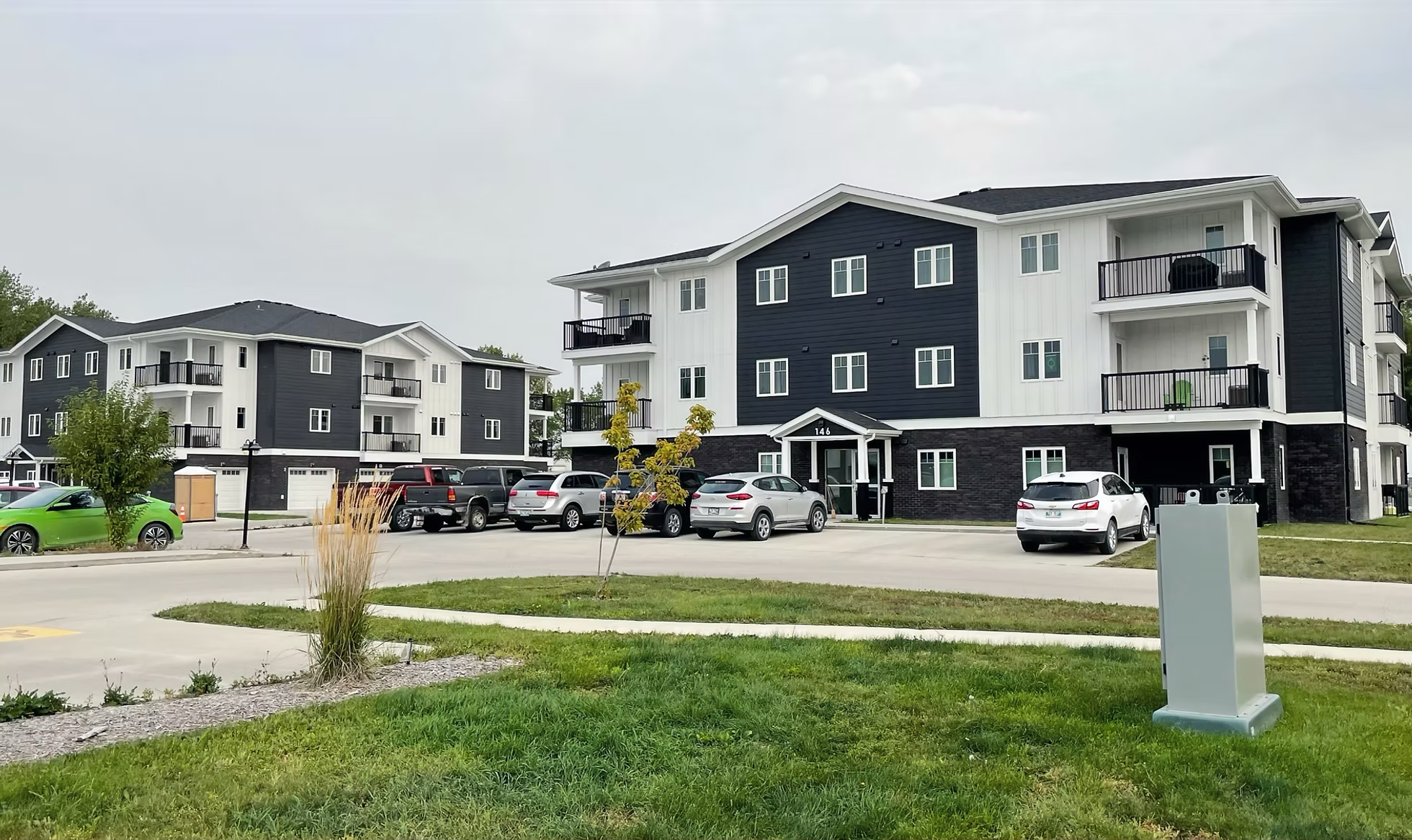 Newly developed apartment complex with three-story buildings in black and white, parked cars in front, and a landscaped area with grass, small trees, and a sidewalk.