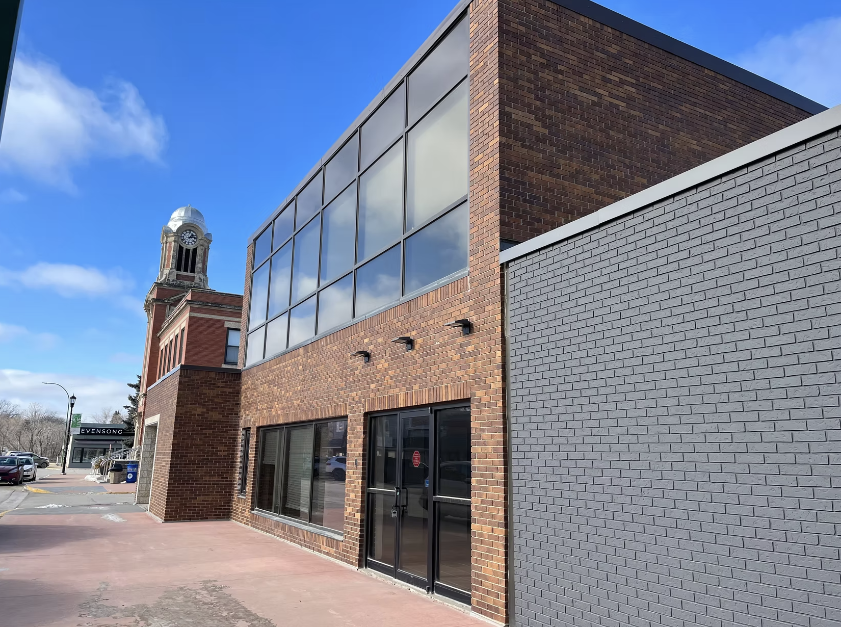Exterior of a modern brick building with large glass windows and a gray brick wall, with a clock tower visible in the background against a partly cloudy blue sky.