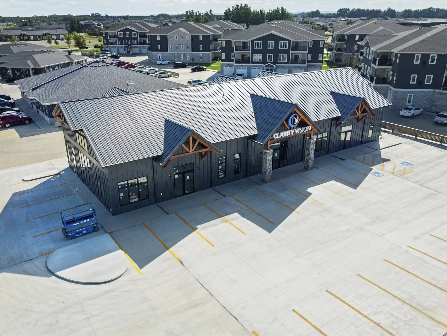 A new commercial building in Winkler with a modern design, dark exterior walls, a metal roof, and the sign 'Clarity Vision' in front of a parking lot with yellow-striped spaces and some handicap spots. Behind are residential houses and trees.