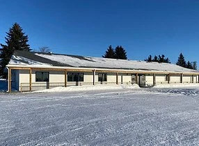A commercial development building with a snow-covered roof, set in a snowy landscape with trees in the background and a clear blue sky overhead.