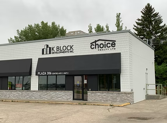 Exterior view of a commercial building development with signs for K Block Developments Inc. and Choice Realty Ltd., showing a black awning over the entrance and a parking lot in front.