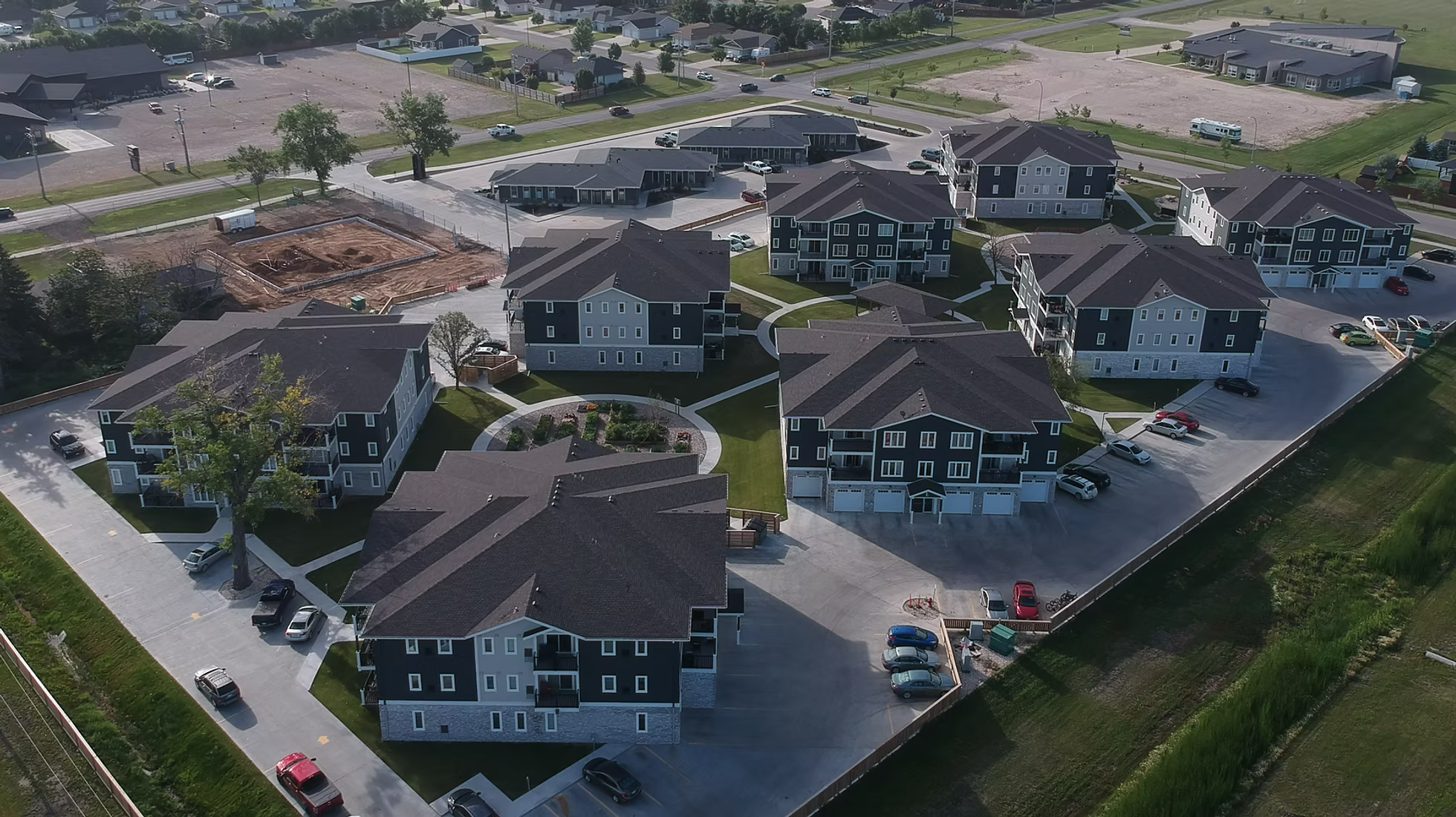 Aerial view of a residential housing development by K Block with multiple apartment buildings, parking lots with cars, green lawns, and trees, with some areas under construction.