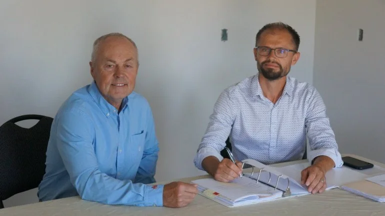 Two men sitting at a table in a room with plain white walls. One man, older with gray hair, wearing a light blue shirt, smiling. The other man, younger with glasses and a beard, wearing a white shirt with small patterns, holding a pen.