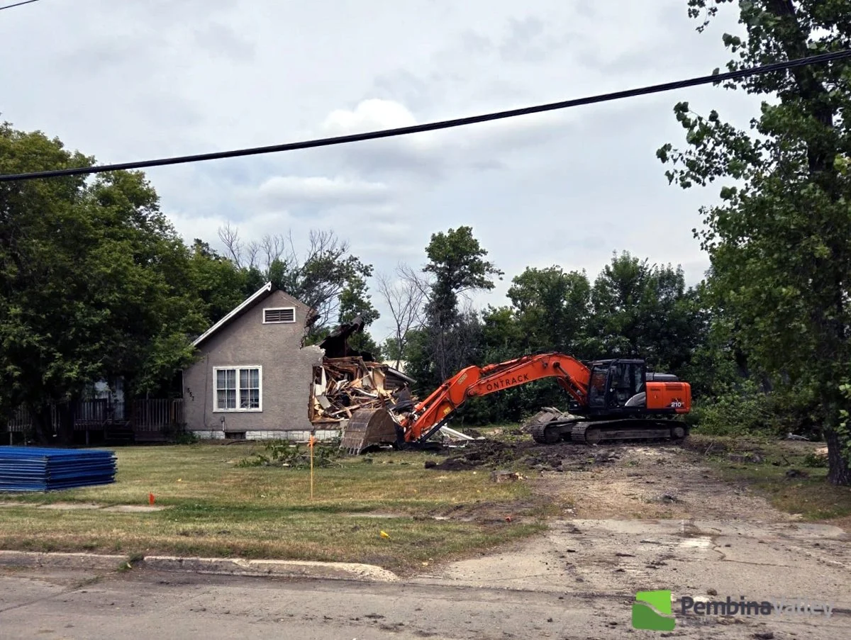 Construction excavator demolishing a house in a residential area with trees and cloudy sky in the background.
