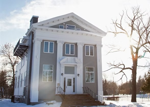 A large, four-story historic house with a prominent front entrance and a symmetrical design, set against a winter landscape with snow, leafless trees, and the sun shining in the background.